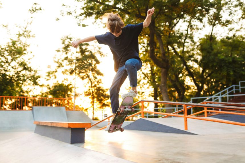 Tânăr patinator cool în tricou negru și blugi care exersează trucuri pe skateboard la skate park