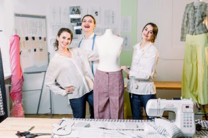 Trei creatori de modă sau croitori zâmbind camerei și măsurând pantaloni lucrați manual pe un manechin într-un atelier confortabil.