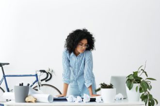 Portret de tânără femeie afro-americană purtând ochelari rotunzi eleganti și cămașă casual, în picioare lângă locul ei de muncă, înconjurată de hârtie rulată, fiind epuizată încercând să găsească o nouă soluție