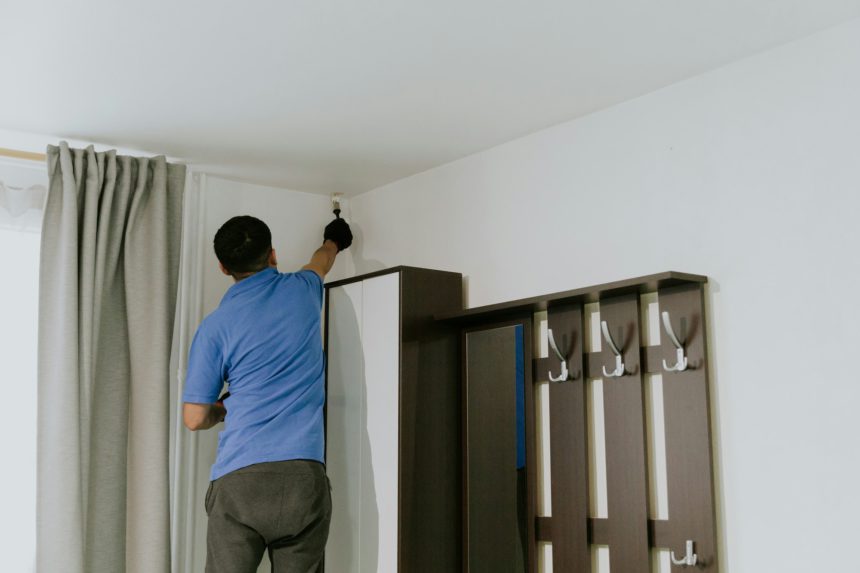 Un tânăr brunet caucazian într-un tricou albastru și pictează colțurile pereților cu vopsea albă folosind o pensulă mică, stând cu spatele pe un scaun lângă fereastră și dulap, vedere laterală în prim-plan cu focalizare selectivă.