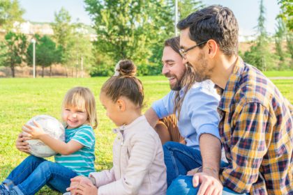 Vedere laterală a unui cuplu de bărbați gay cu cei doi copii ai lor stând pe iarbă într-un parc distrându-se în familie.