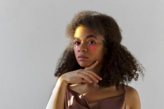 Portret de studio creativ al tânărului model afro-american creț cu machiaj artistic de culoare strălucitoare uitându-se la camera, femeie de culoare cu vopsea roz pe jumătatea feței pozând izolat pe fundal gri