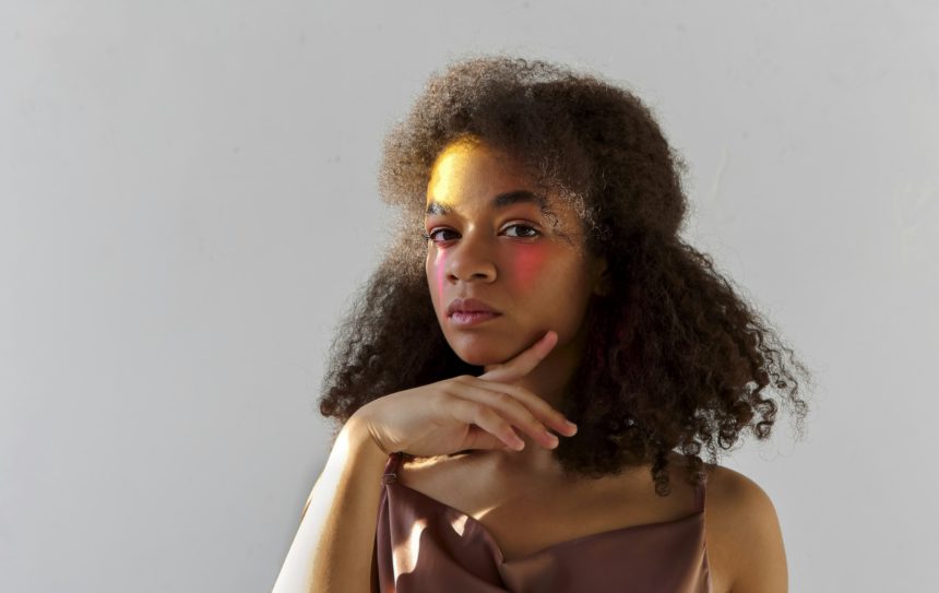 Portret de studio creativ al tânărului model afro-american creț cu machiaj artistic de culoare strălucitoare uitându-se la camera, femeie de culoare cu vopsea roz pe jumătatea feței pozând izolat pe fundal gri