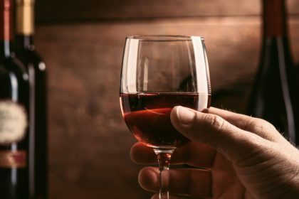 Experiență de degustare de vinuri în pivnița rustică și barul de vinuri: somelier ținând un pahar de vin roșu delicios și o colecție excelentă de sticle de vin pe fundal