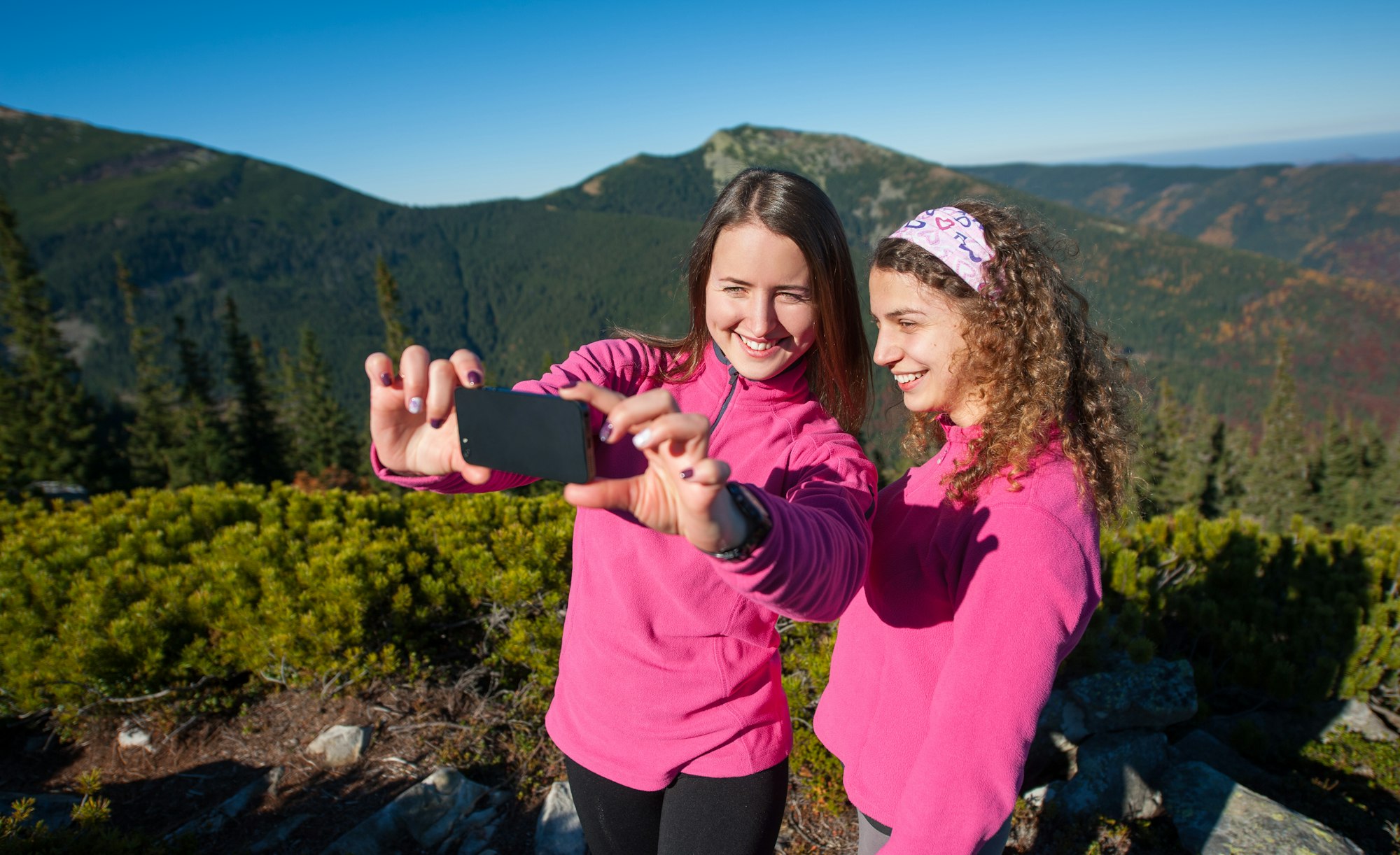 Două excursioniste fericite zâmbind și făcând selfie-uri folosind telefonul mobil în vârful stâncii de munte.