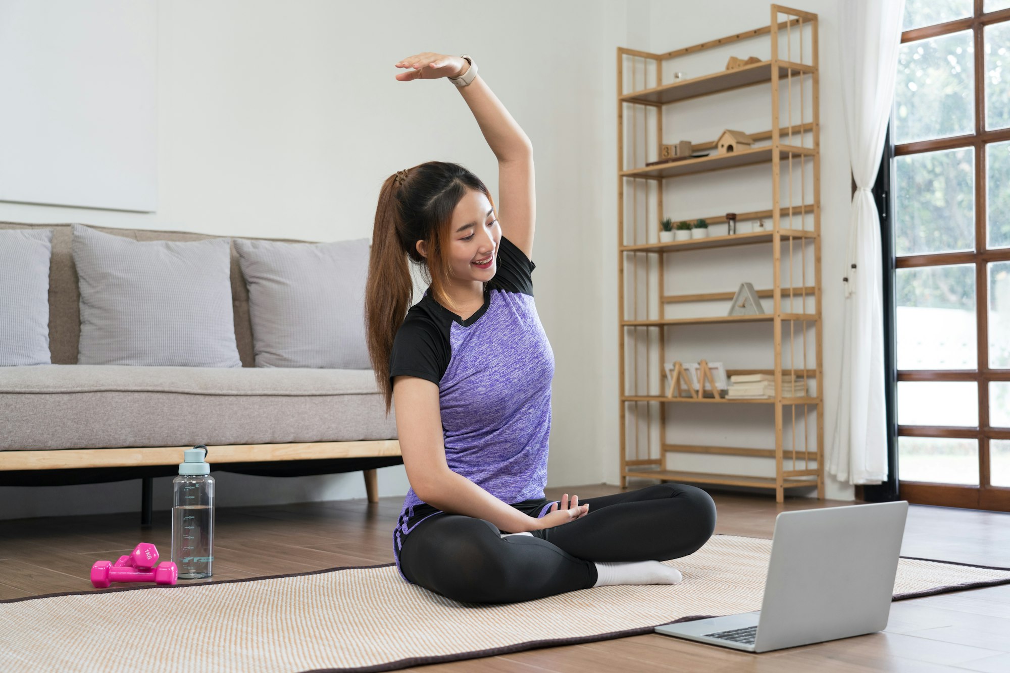 Tânără femeie asiatică care urmărește videoclipuri de fitness pe laptop pentru a se antrena și a face exerciții pentru a întinde brațul.