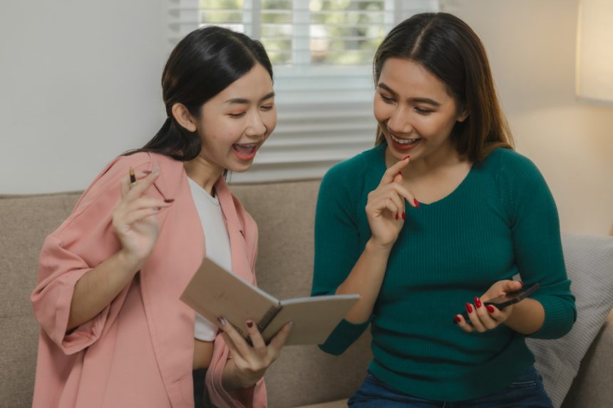 Două femei frumoase din Asia folosesc dispozitive electronice pentru a se distra ca o activitate de grup în apartamentul lor, petrec timpul liber împreună pe canapea de acasă în weekend.