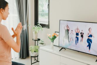 Tânără femeie asiatică atractivă în site-ul de îmbrăcăminte sport pe covoraș în podea clasă virtuală de fitness cu un grup de oameni la o conferință video la televizor practică yoga pentru începători în camera de zi acasă.