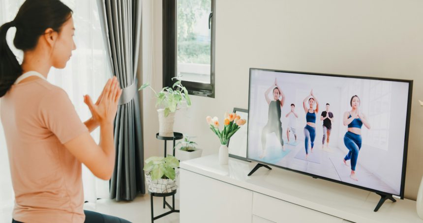 Tânără femeie asiatică atractivă în site-ul de îmbrăcăminte sport pe covoraș în podea clasă virtuală de fitness cu un grup de oameni la o conferință video la televizor practică yoga pentru începători în camera de zi acasă.
