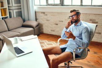 Un bărbat în cămașă și pantaloni scurți stă la o masă și vorbește la un apel video, o conferință de afaceri online, o întâlnire.