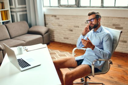 Un bărbat în cămașă și pantaloni scurți stă la o masă și vorbește la un apel video, o conferință de afaceri online, o întâlnire.
