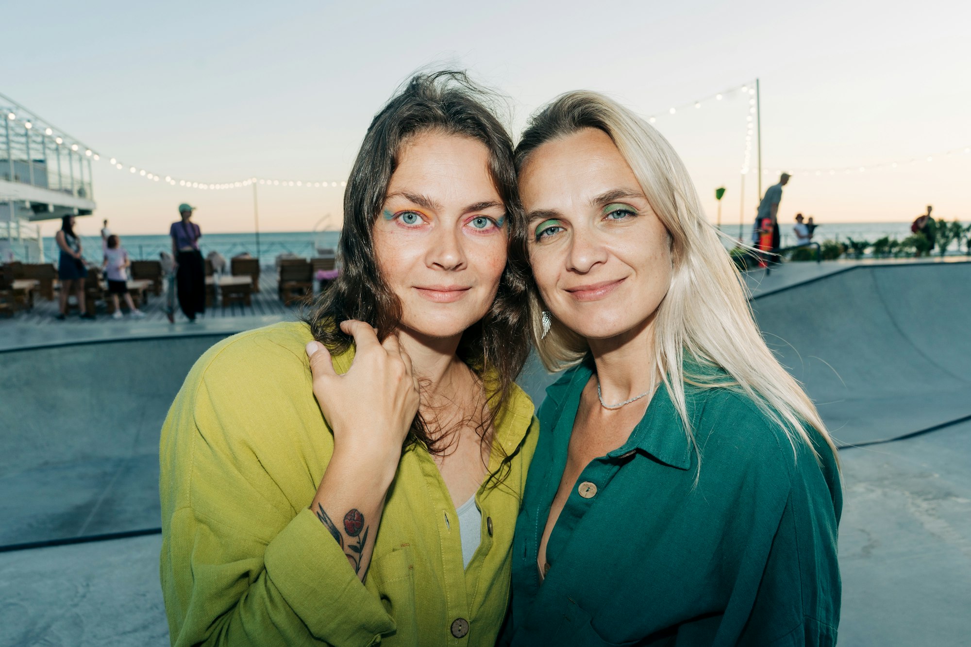 Două femei frumoase cu machiaj multicolor pe ochi se uită la aparatul de fotografiat în timp ce stau pe stradă.