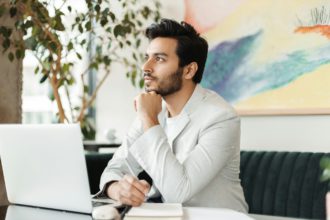 Tânăr profesionist care se gândește la strategia de afaceri, luând note în timp ce lucrează la laptop într-un spațiu de birou elegant