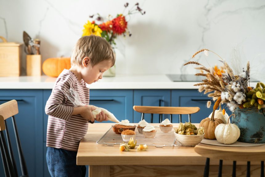 Băiat drăguț care decorează cupcakes cu dovleac cu cremă de brânză și cireșe de iarnă în bucătăria scandinavă