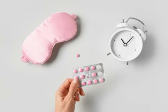 Mască de dormit roz, ceas, alarmă și capsule de vitamine roz pe fundal gri.