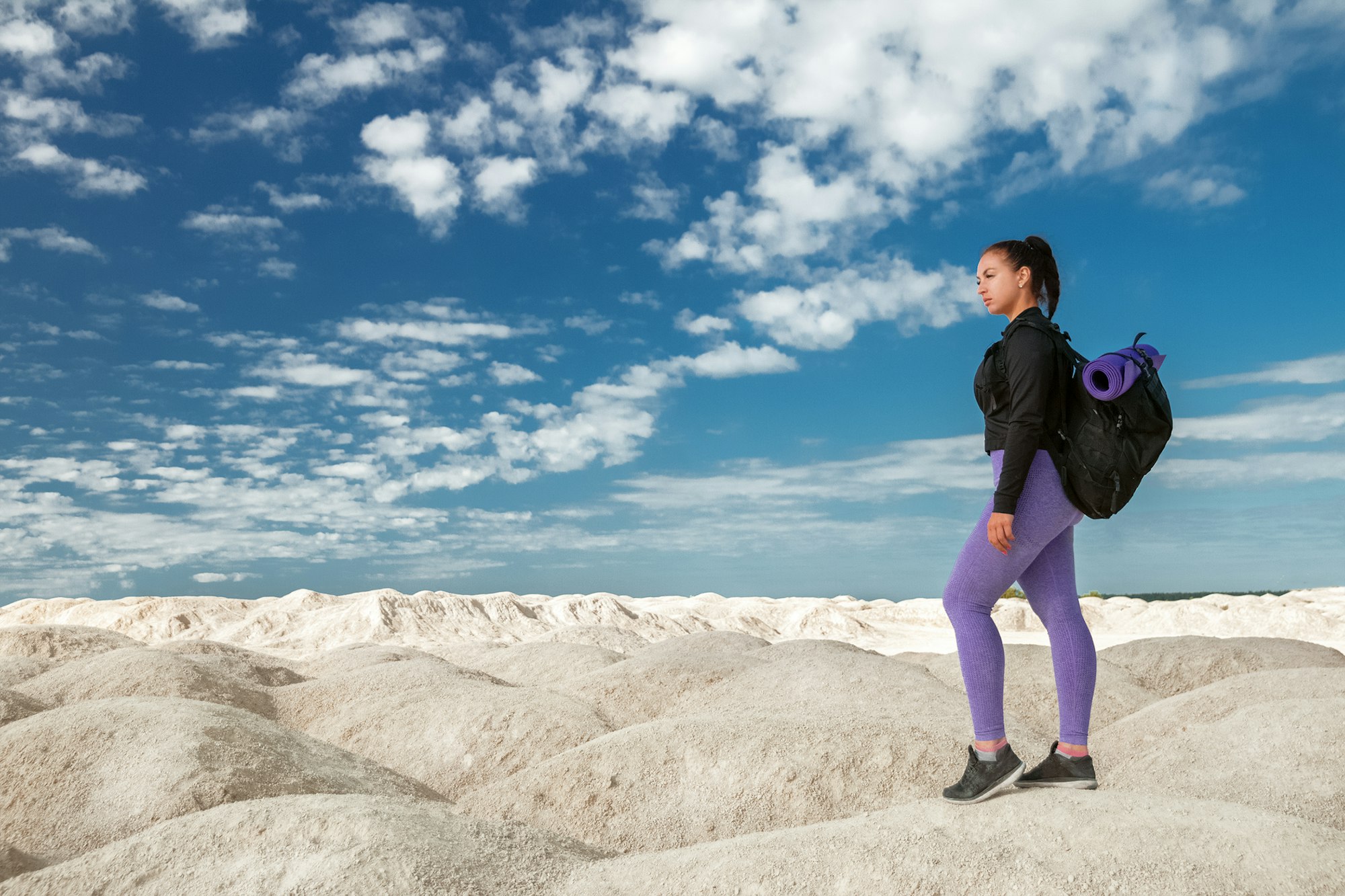 Tânără sportivă excursionist solo în îmbrăcăminte sport și cu rucsac ținând sport arată spre stânga pe deșertul deluros cu cer albastru înnorat, spațiu pentru copiere