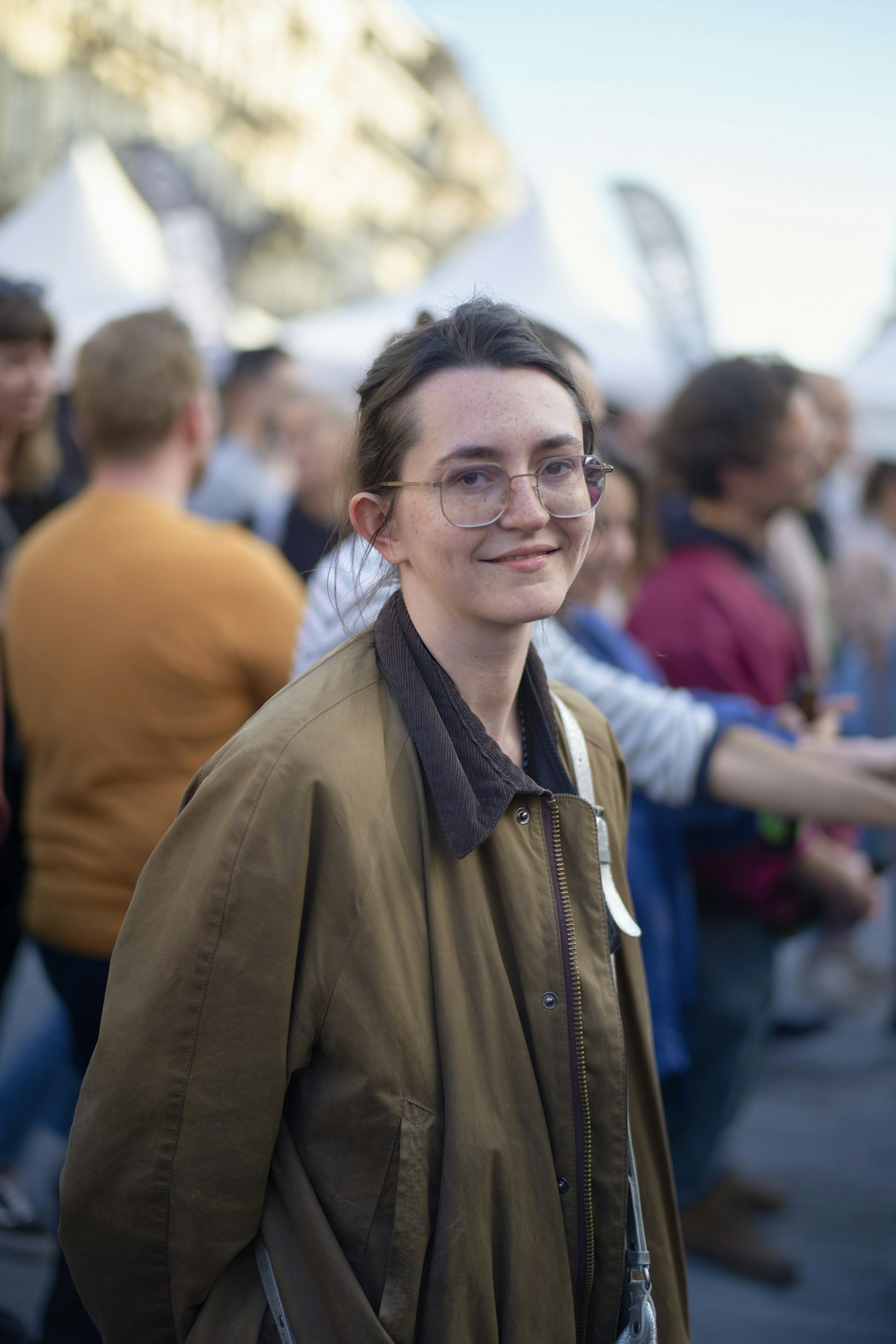 Femeie zâmbitoare purtând ochelari cu o mulțime în fundal în timpul unui eveniment în aer liber.