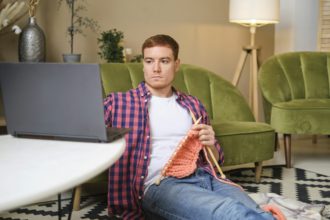 tânăr cu părul roșu, îmbrăcat într-o cămașă roșie, studiază cu atenție priceperea de a tricota în timp ce urmărește un videoclip cu instrucțiuni pe computerul său.