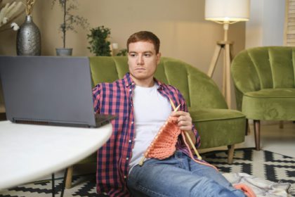 tânăr cu părul roșu, îmbrăcat într-o cămașă roșie, studiază cu atenție priceperea de a tricota în timp ce urmărește un videoclip cu instrucțiuni pe computerul său.