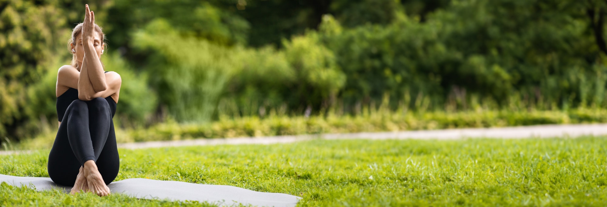 Doamnă flexibilă care practică yoga într-un parc public, femeie în îmbrăcăminte sport neagră stând pe saltea de fitness cu brațele și picioarele încrucișate, lucrând la puterea corpului, panoramă cu spațiu de copiere